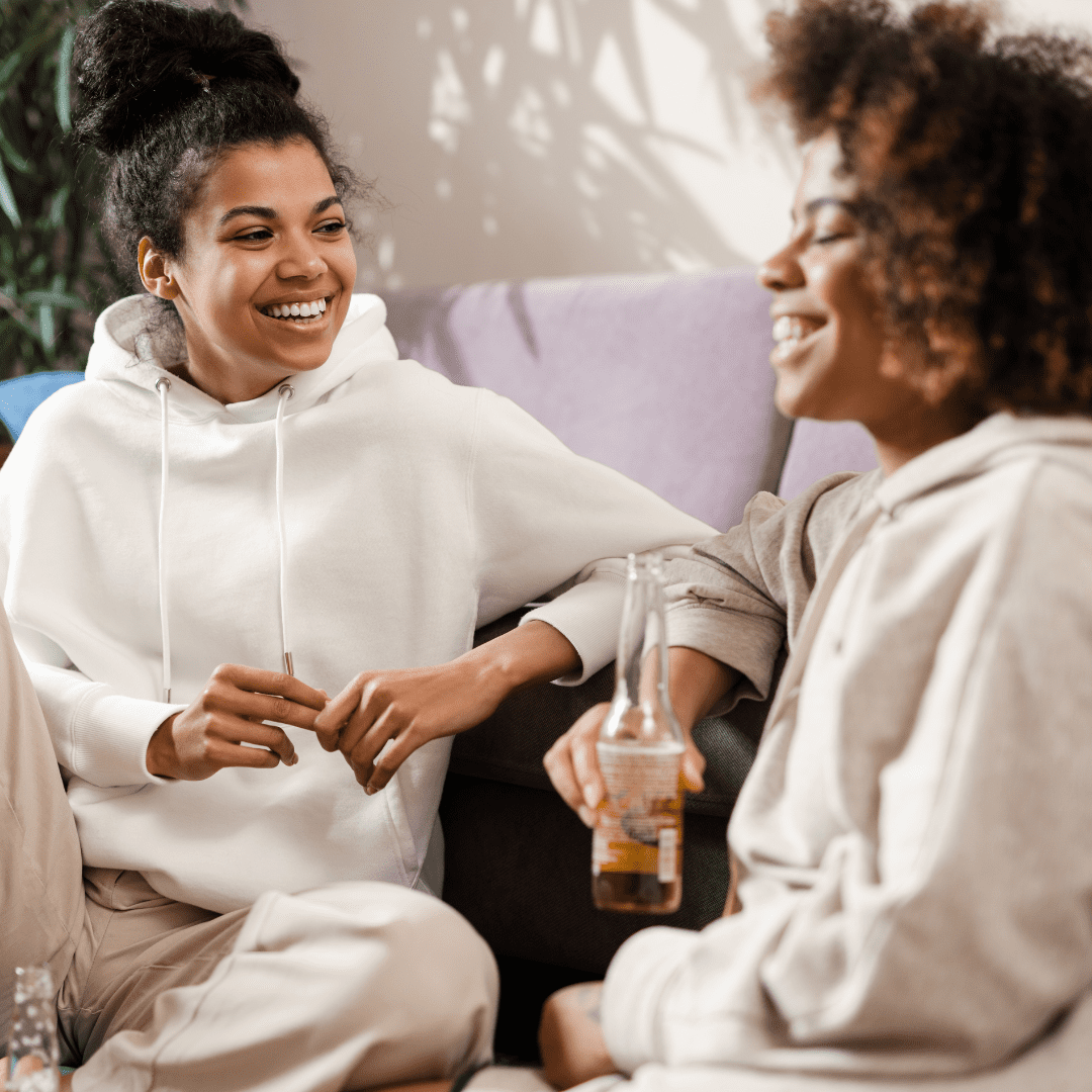 Black women laughing and talking in a home environment with drinks in their hand | How to stop being a people pleaser