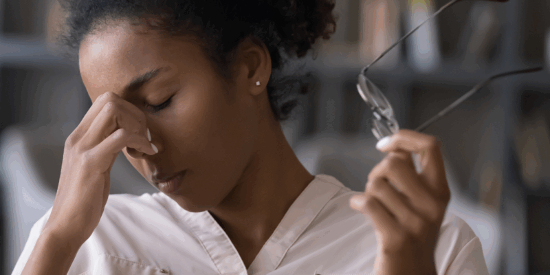 Black woman sitting at desk in front of laptop, massaging the bridge of her nose, with eyeglasses in the other hand. | How can I stop overthinking?