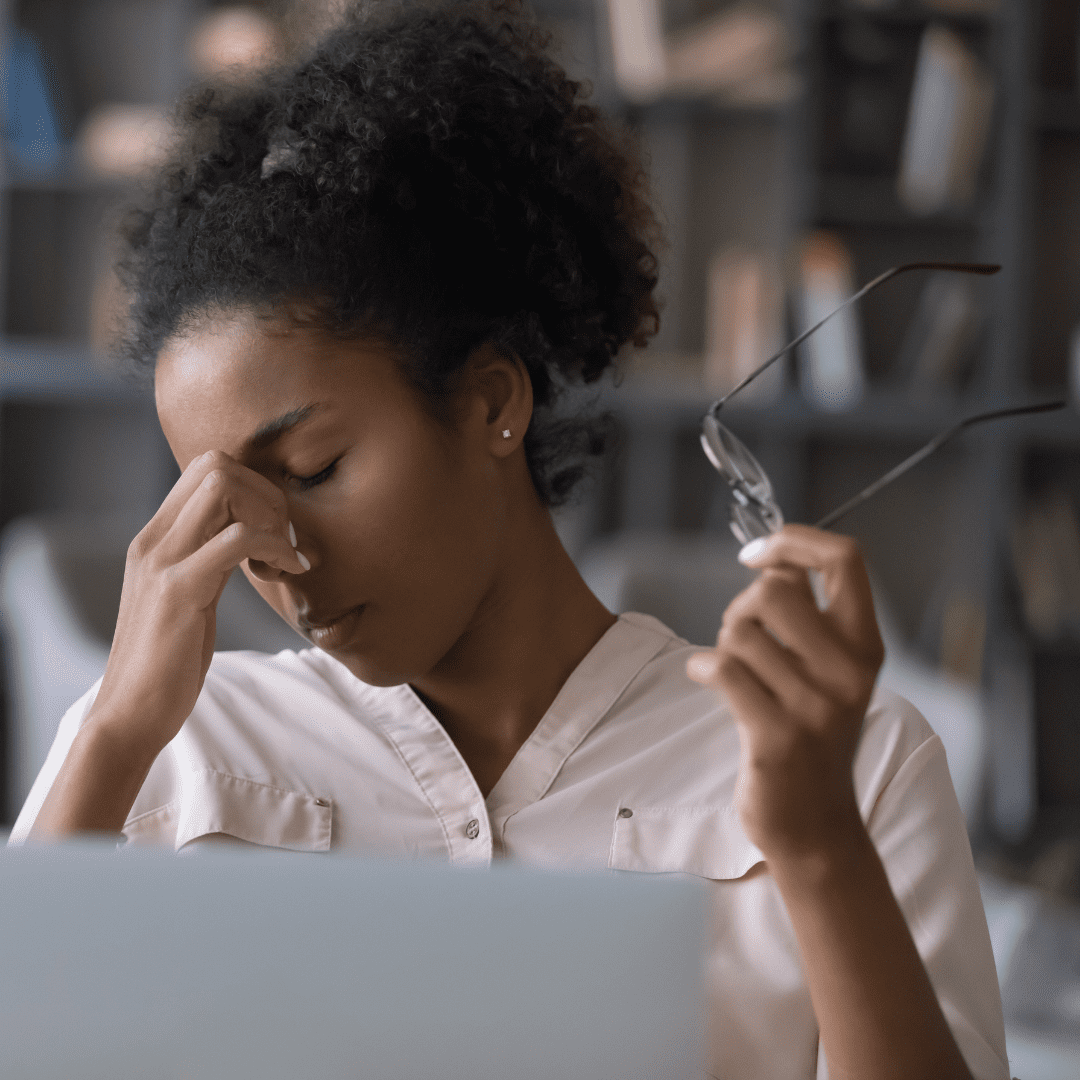 Black woman sitting at desk in front of laptop, massaging the bridge of her nose, with eyeglasses in the other hand. | How can I stop overthinking?