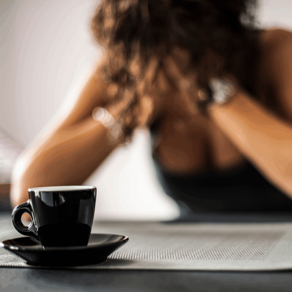 Black woman sitting at table with head in hands with a cup of coffee in front of her | Why do I feel anxious when I wake up