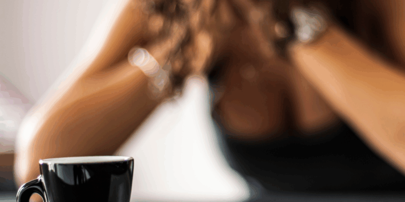 Black woman sitting at table with head in hands with a cup of coffee in front of her | Why do I feel anxious when I wake up