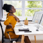 Black woman sitting at work desk with one hand on neck and another on lower back | how emotions are stored in the body